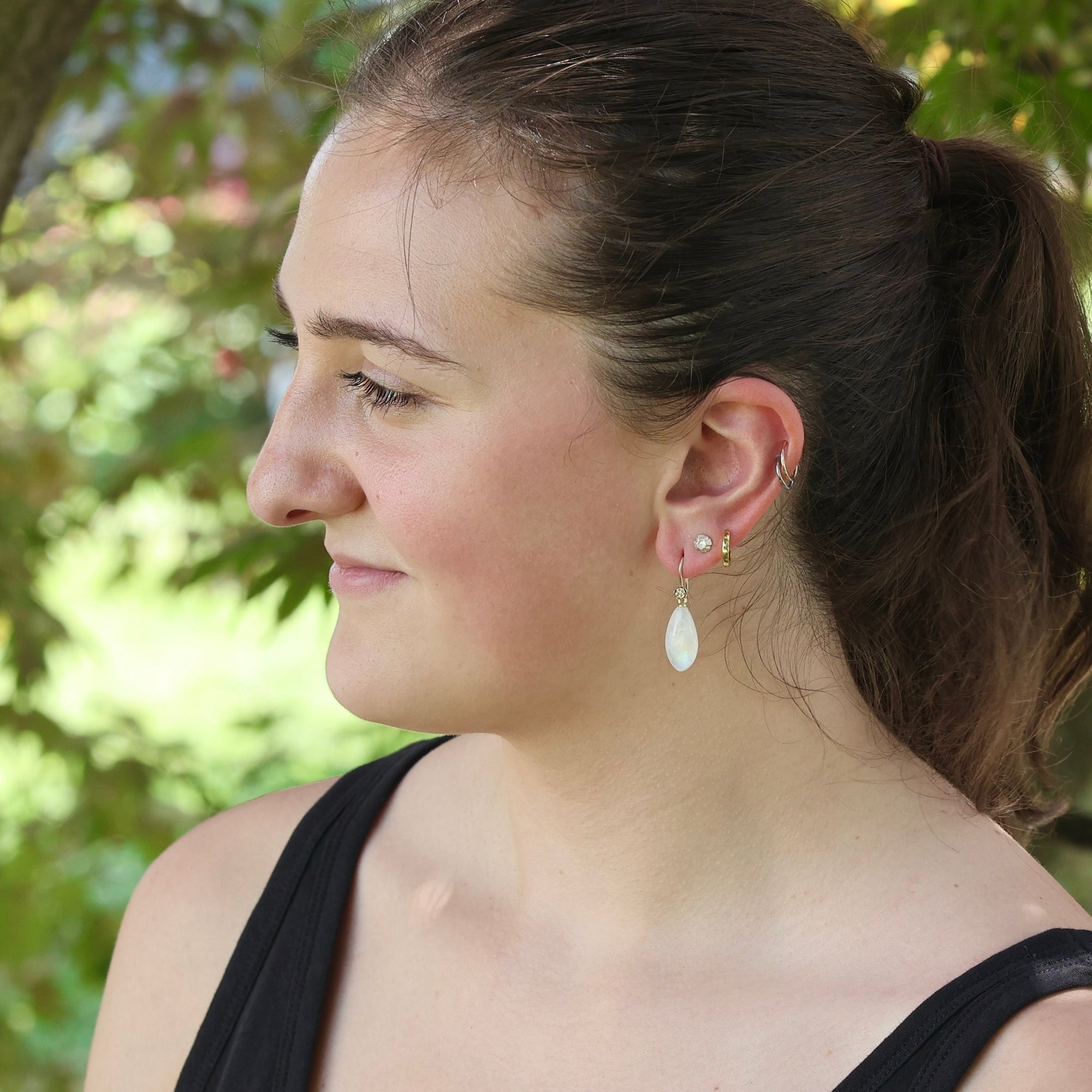A woman in profile wearing a small white teardrop dangle earring with gold-toned posts, photographed outdoors with a blurred green background.