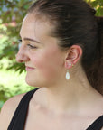 A woman in profile wearing a small white teardrop dangle earring with gold-toned posts, photographed outdoors with a blurred green background.