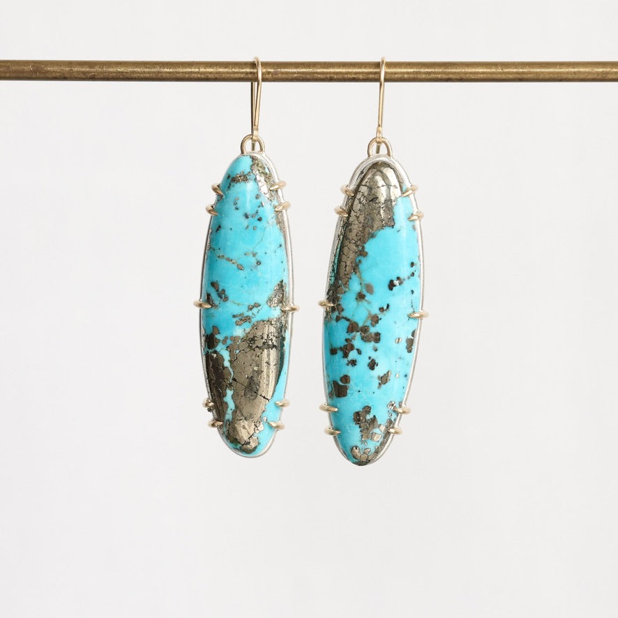 A pair of long oval drop earrings with bright turquoise-blue marbled stones and dark matrix, set in gold-toned metal and hanging from gold hooks against a plain light background.