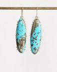 A pair of long oval drop earrings with bright turquoise-blue marbled stones and dark matrix, set in gold-toned metal and hanging from gold hooks against a plain light background.