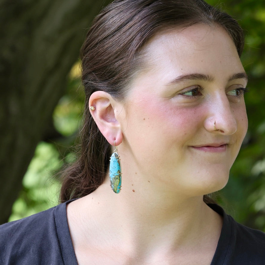 A woman wearing a long turquoise and brown stone dangle earring, photographed outdoors with a blurred green and brown natural background.