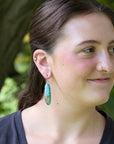A woman wearing a long turquoise and brown stone dangle earring, photographed outdoors with a blurred green and brown natural background.