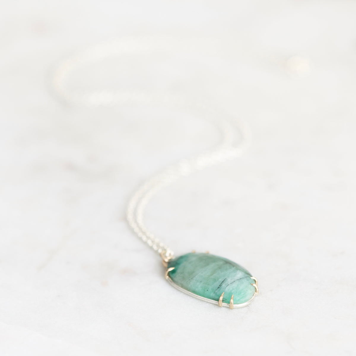 A silver chain necklace with an oval green stone pendant in a gold-toned bezel setting, resting on a light marble surface.