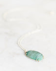 A silver chain necklace with an oval green stone pendant in a gold-toned bezel setting, resting on a light marble surface.