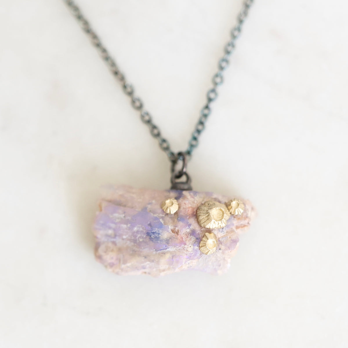A fossilized opal adorned with gold barnacle details set east west on an oxidized silver chain laid out on a marble background. 