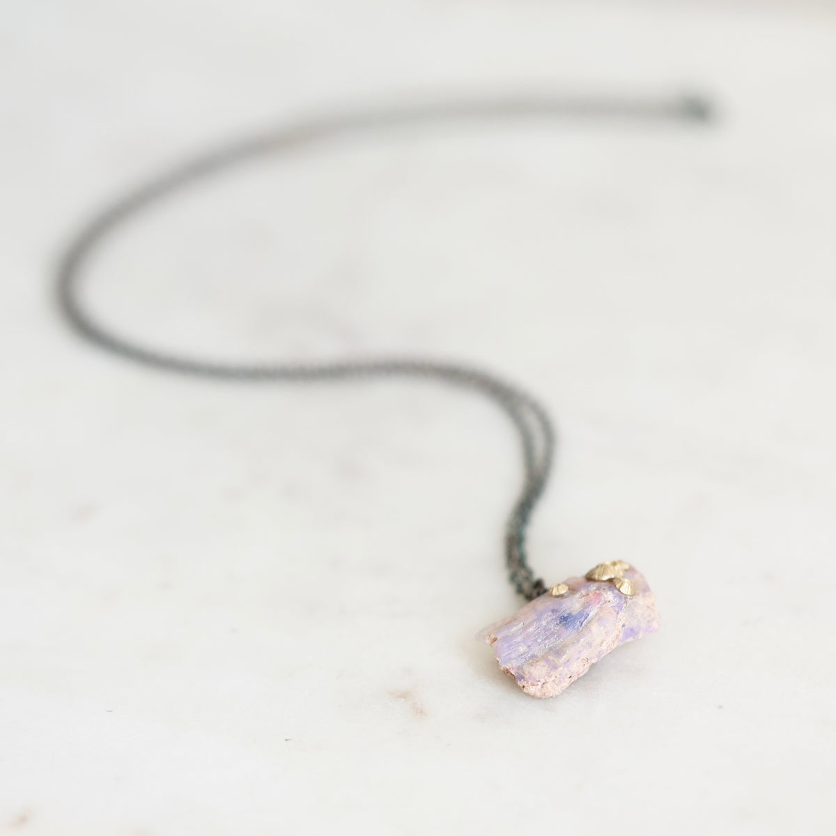 A fossilized opal adorned with gold barnacle details set east west on an oxidized silver chain laid out on a marble background. 