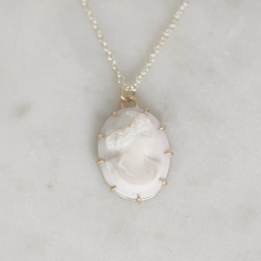 A delicate necklace featuring a classic oval cameo pendant with a carved profile of a woman, set in a gold-toned bezel with small gold accent prongs. The pendant hangs from a fine gold chain against a light gray marble background.