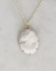 A delicate necklace featuring a classic oval cameo pendant with a carved profile of a woman, set in a gold-toned bezel with small gold accent prongs. The pendant hangs from a fine gold chain against a light gray marble background.