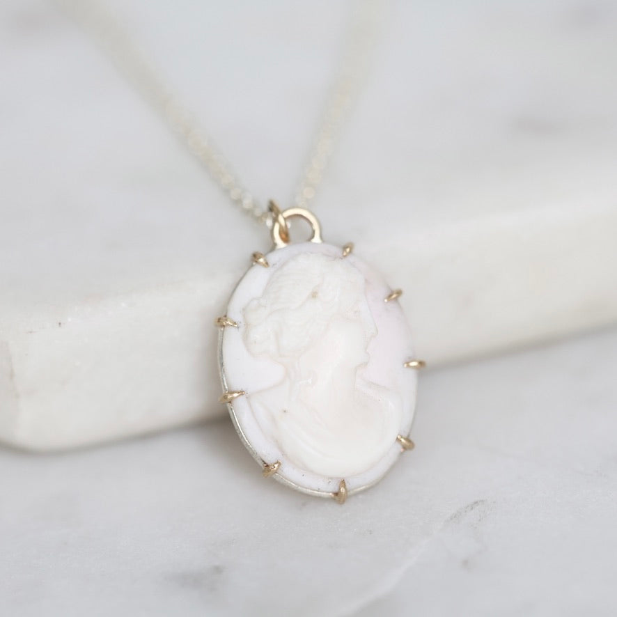 A close-up of a vintage-style oval pendant necklace featuring a carved white cameo face in a gold-toned bezel, resting on a white marble block against a light marble surface.