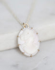 A close-up of a vintage-style oval pendant necklace featuring a carved white cameo face in a gold-toned bezel, resting on a white marble block against a light marble surface.