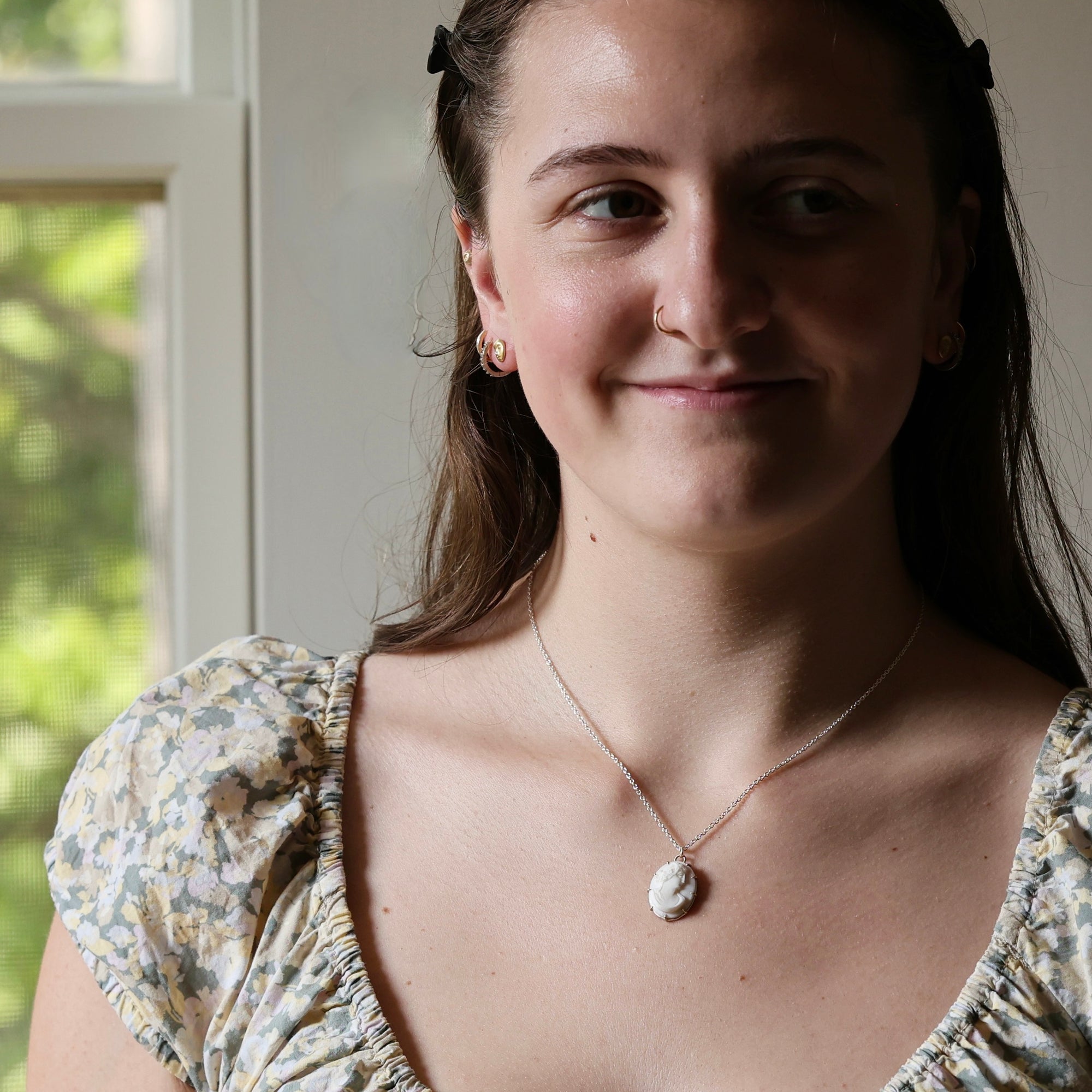 A young woman with brown hair and a nose ring wearing a delicate silver necklace with a white shell pendant, paired with a floral puff-sleeve top, photographed in a bright indoor setting.