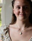 A young woman with brown hair and a nose ring wearing a delicate silver necklace with a white shell pendant, paired with a floral puff-sleeve top, photographed in a bright indoor setting.