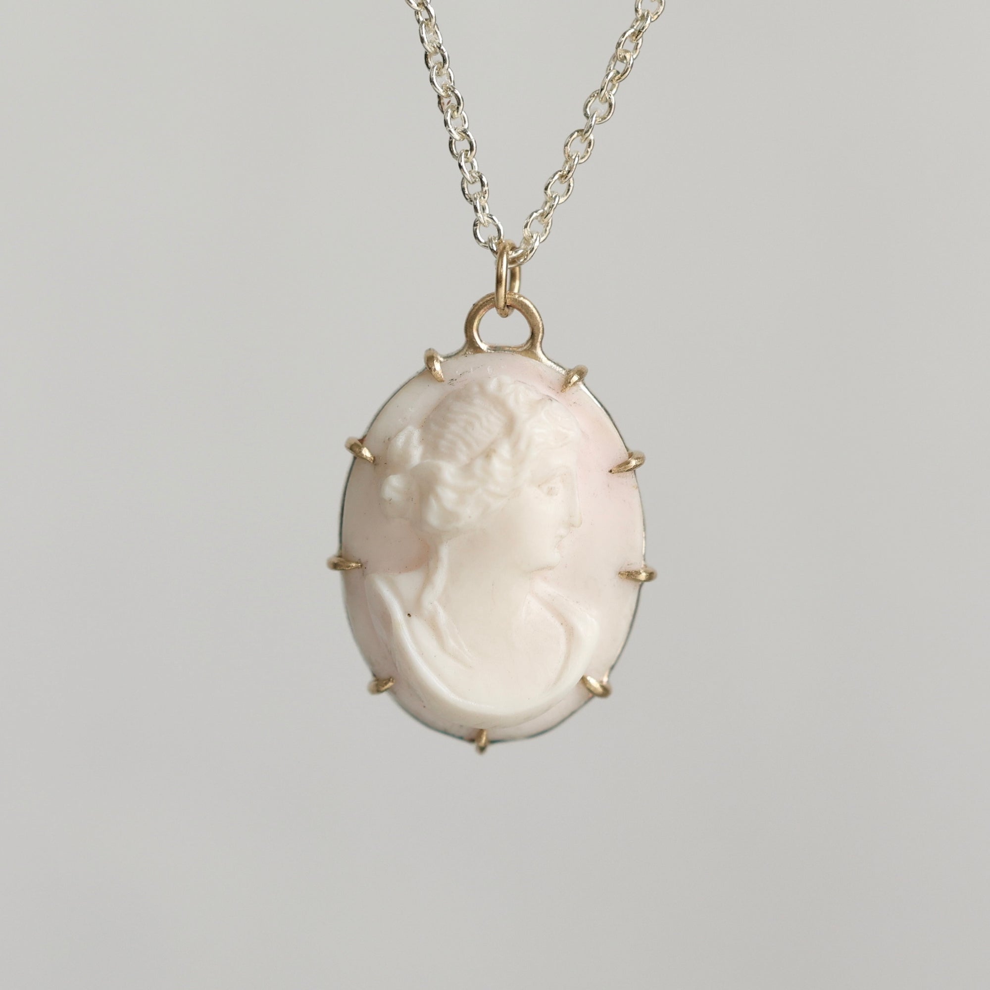 A vintage-style oval pendant necklace featuring a pale pink carved cameo profile in a gold-toned bezel with small spike accents, suspended from a delicate gold chain against a neutral gray background.