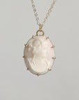 A vintage-style oval pendant necklace featuring a pale pink carved cameo profile in a gold-toned bezel with small spike accents, suspended from a delicate gold chain against a neutral gray background.