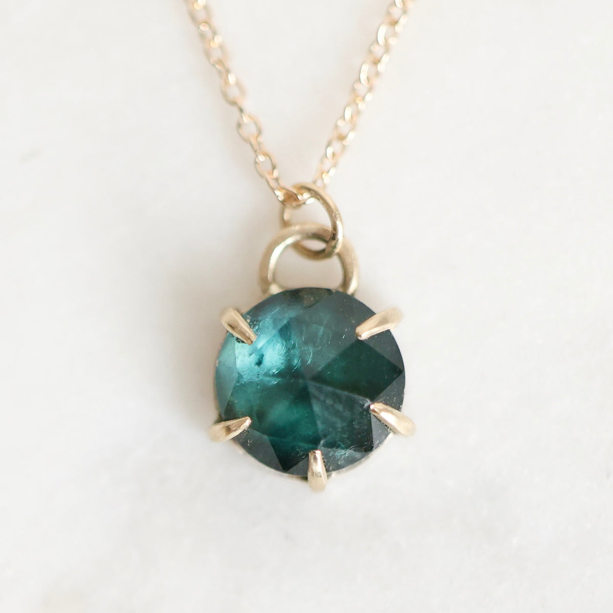 A gold necklace with a faceted round dark teal gemstone pendant in a prong setting, shown on a light off-white background.