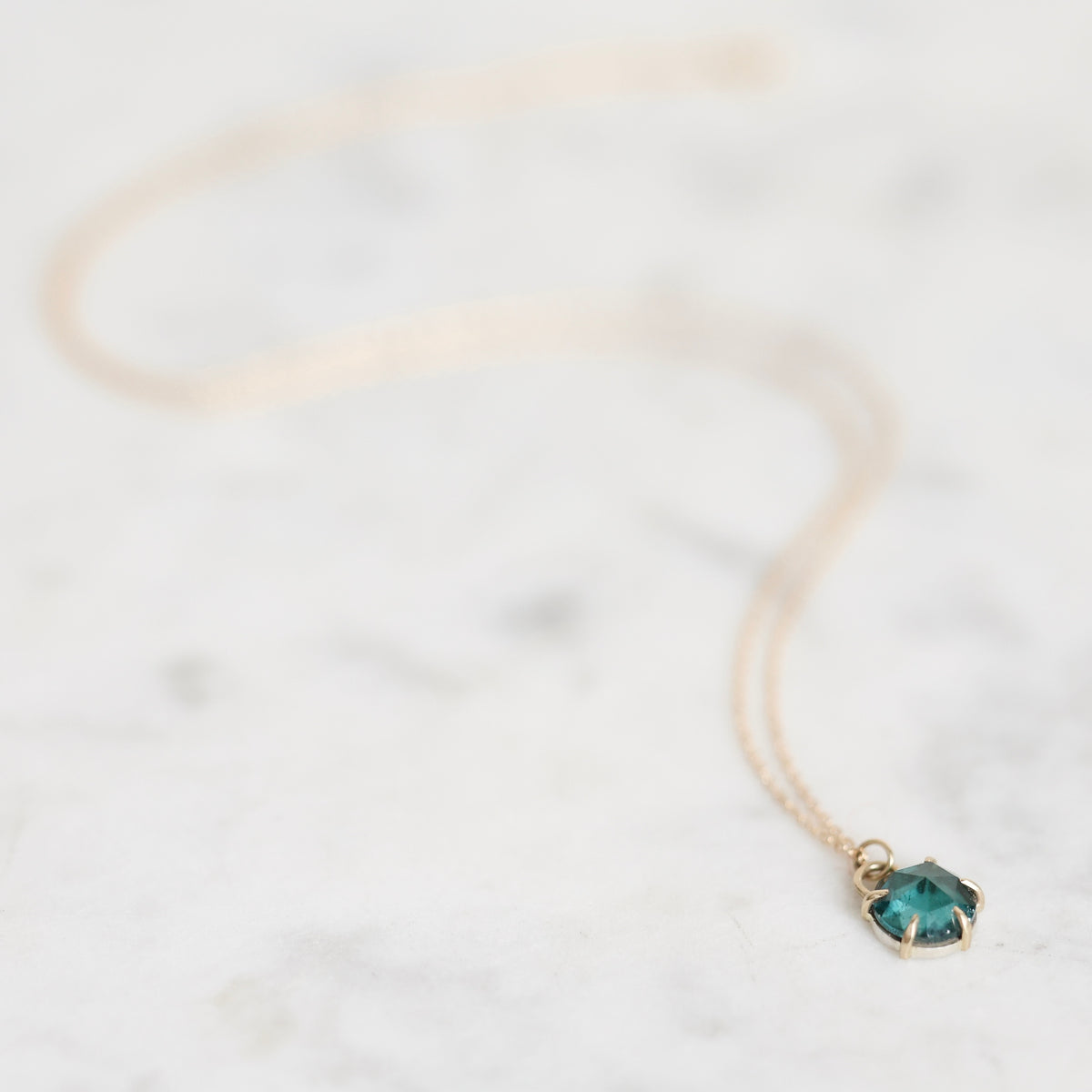 A delicate gold chain necklace with a small, faceted teal-green gemstone pendant in a gold bezel setting, resting on a light marble surface.
