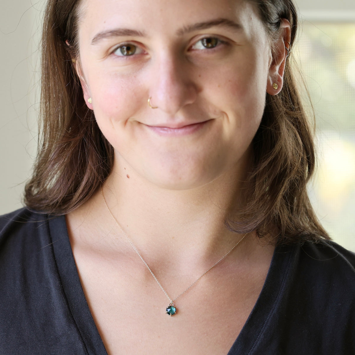 A woman with brown hair wearing a delicate chain necklace with a small round teal gemstone pendant, shown in a close-up portrait.
