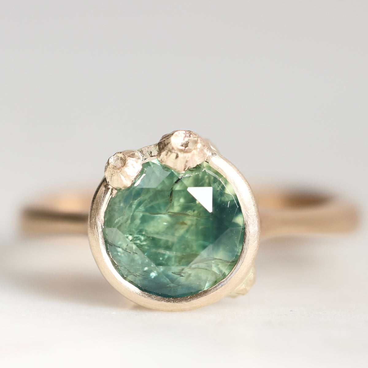 A close-up shot of a gold ring featuring a round, faceted green gemstone with natural inclusions, set in a textured gold bezel with small accent stones on top, on a light gray background.