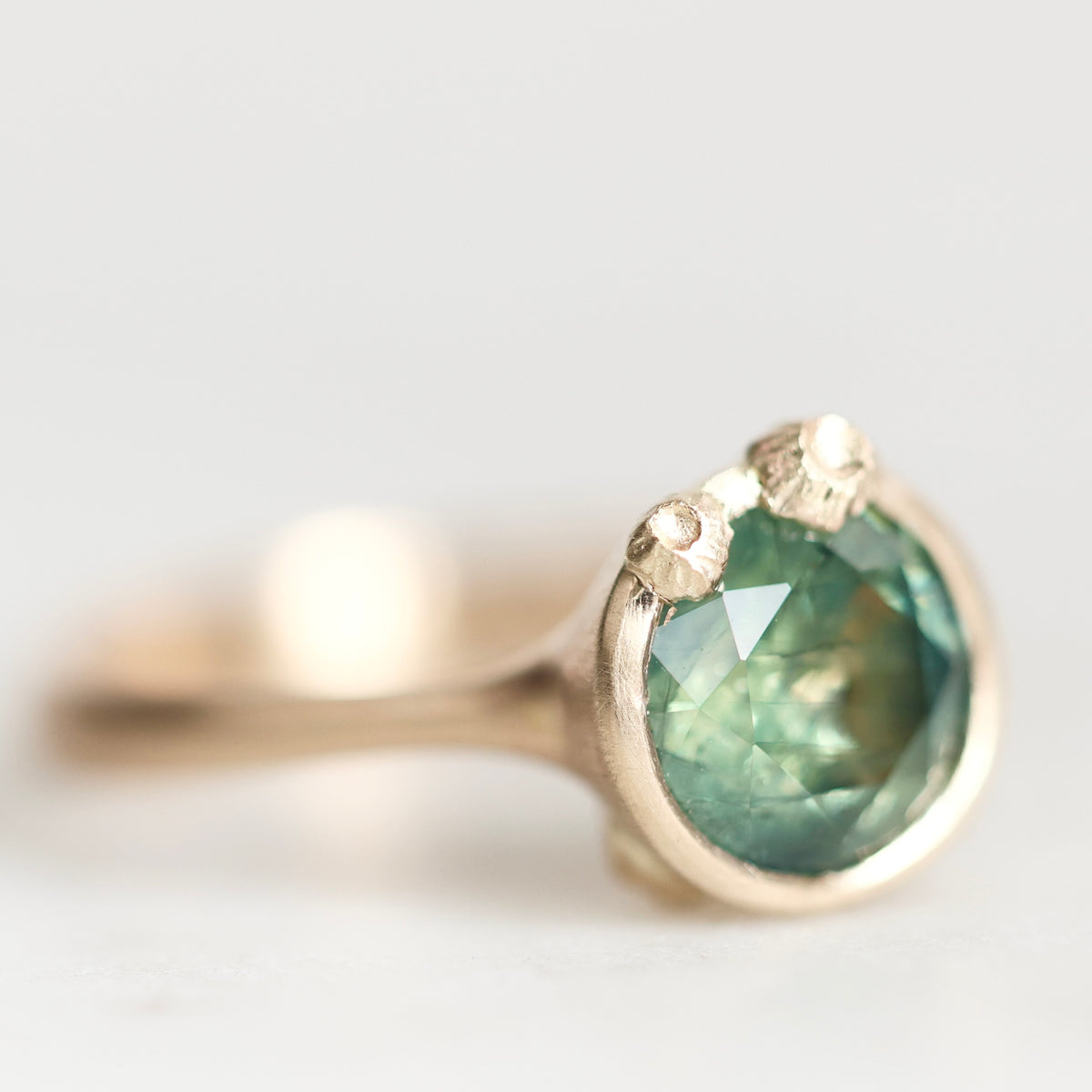 A close-up shot of a gold ring featuring a round faceted green gemstone in a prong setting, with two small accent stones on the shoulders, set against a soft, out-of-focus light gray background.