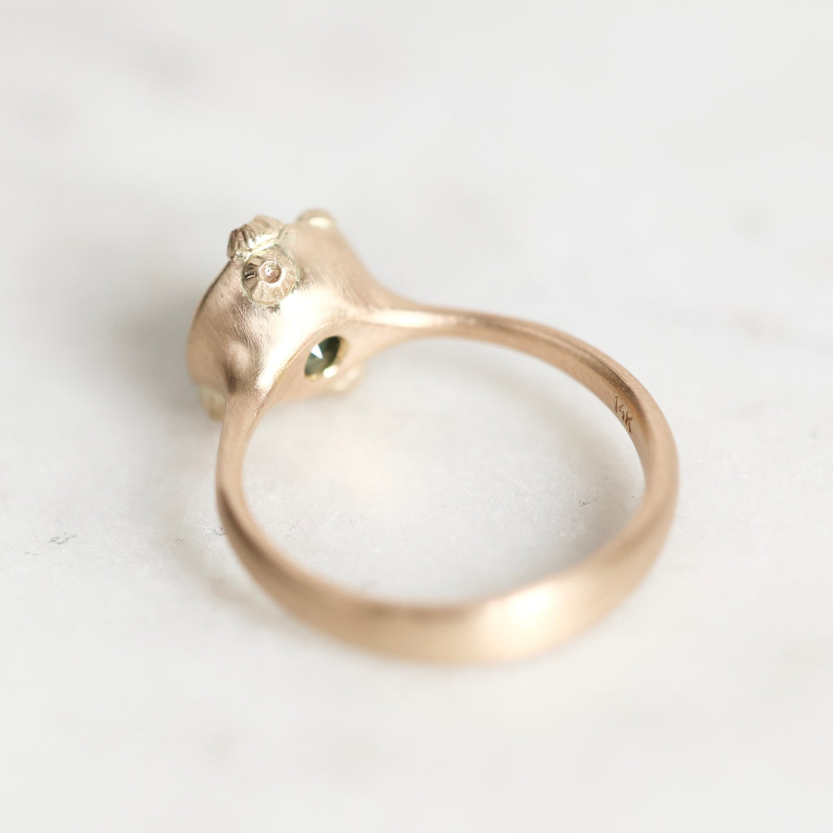 A close-up of a gold ring with a sculptural, organic-shaped top featuring a tiny green stone, resting on a light marble surface.