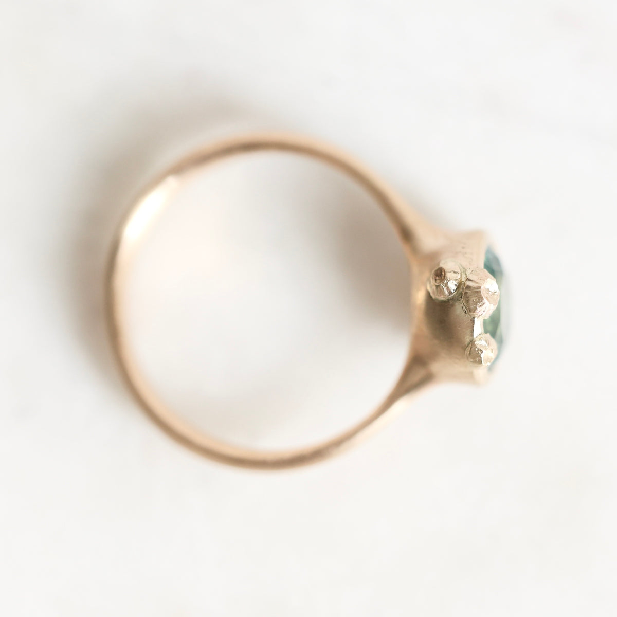 A close-up shot of a gold ring with a small green faceted gemstone in a prong setting, photographed on a light, neutral background.