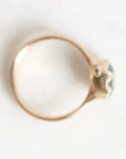 A close-up shot of a gold ring with a small green faceted gemstone in a prong setting, photographed on a light, neutral background.