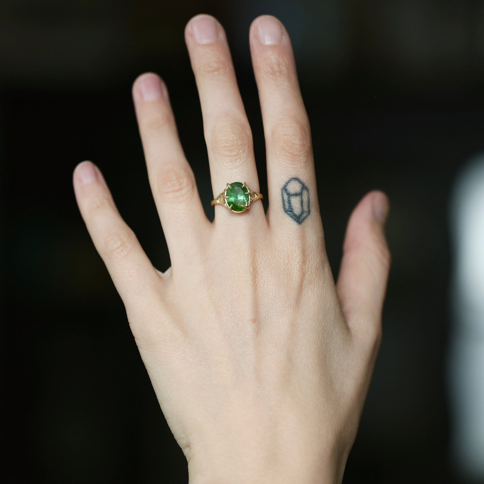 A close-up of a hand wearing a gold ring with a green oval gemstone, set against a dark blurred background.