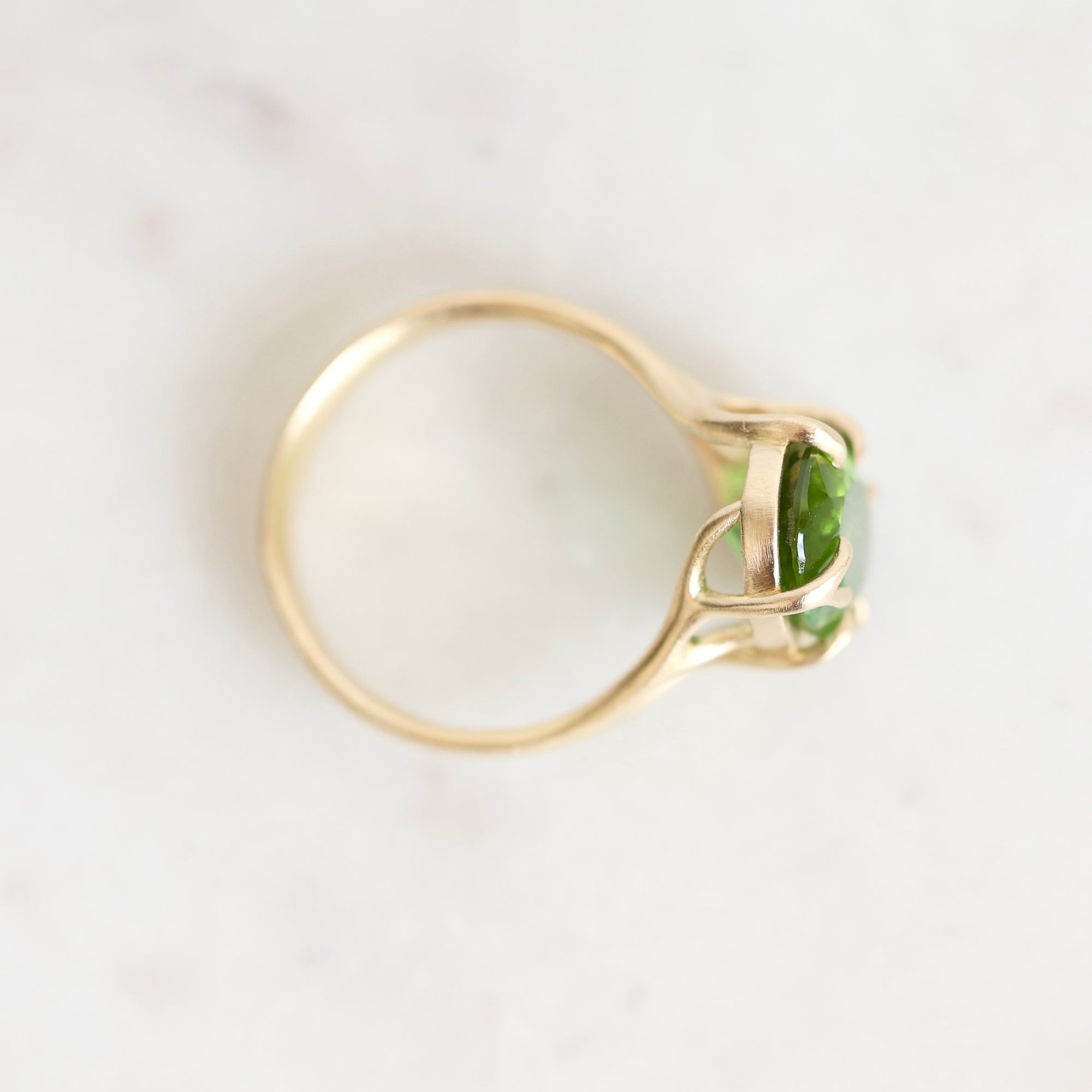 A yellow gold ring with a marquise-cut green gemstone, photographed from a top-down angle on a light, off-white marble surface.
