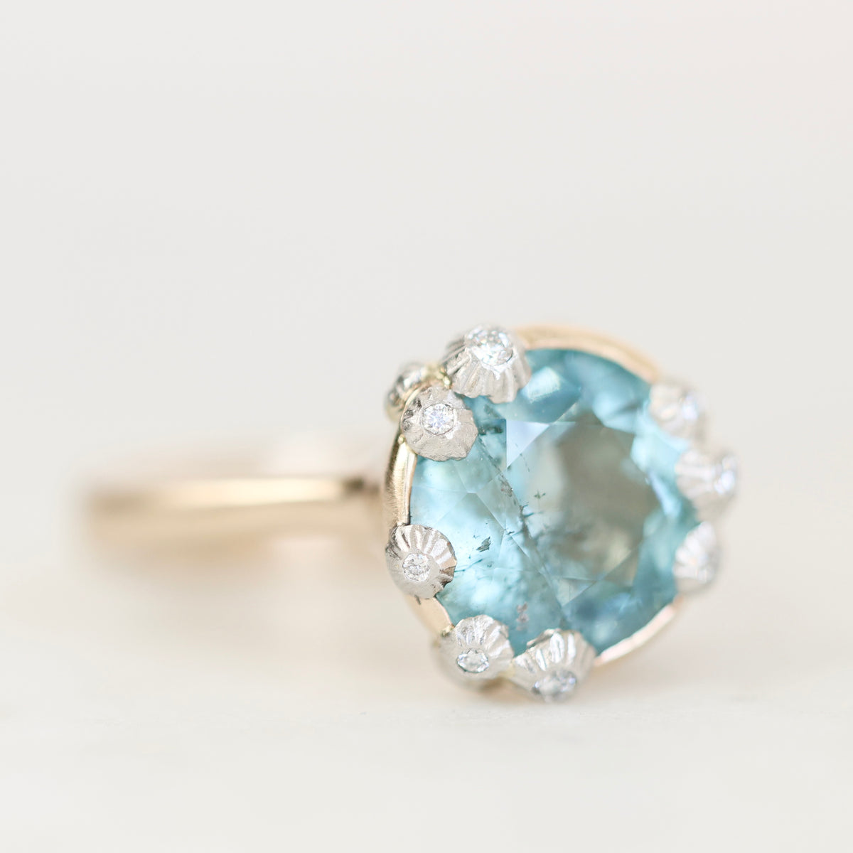 A close-up shot of a ring featuring a large oval-cut light blue gemstone surrounded by small clear accent stones in a silver-toned setting, on a plain light background.