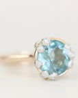 A close-up shot of a ring featuring a large oval-cut light blue gemstone surrounded by small clear accent stones in a silver-toned setting, on a plain light background.