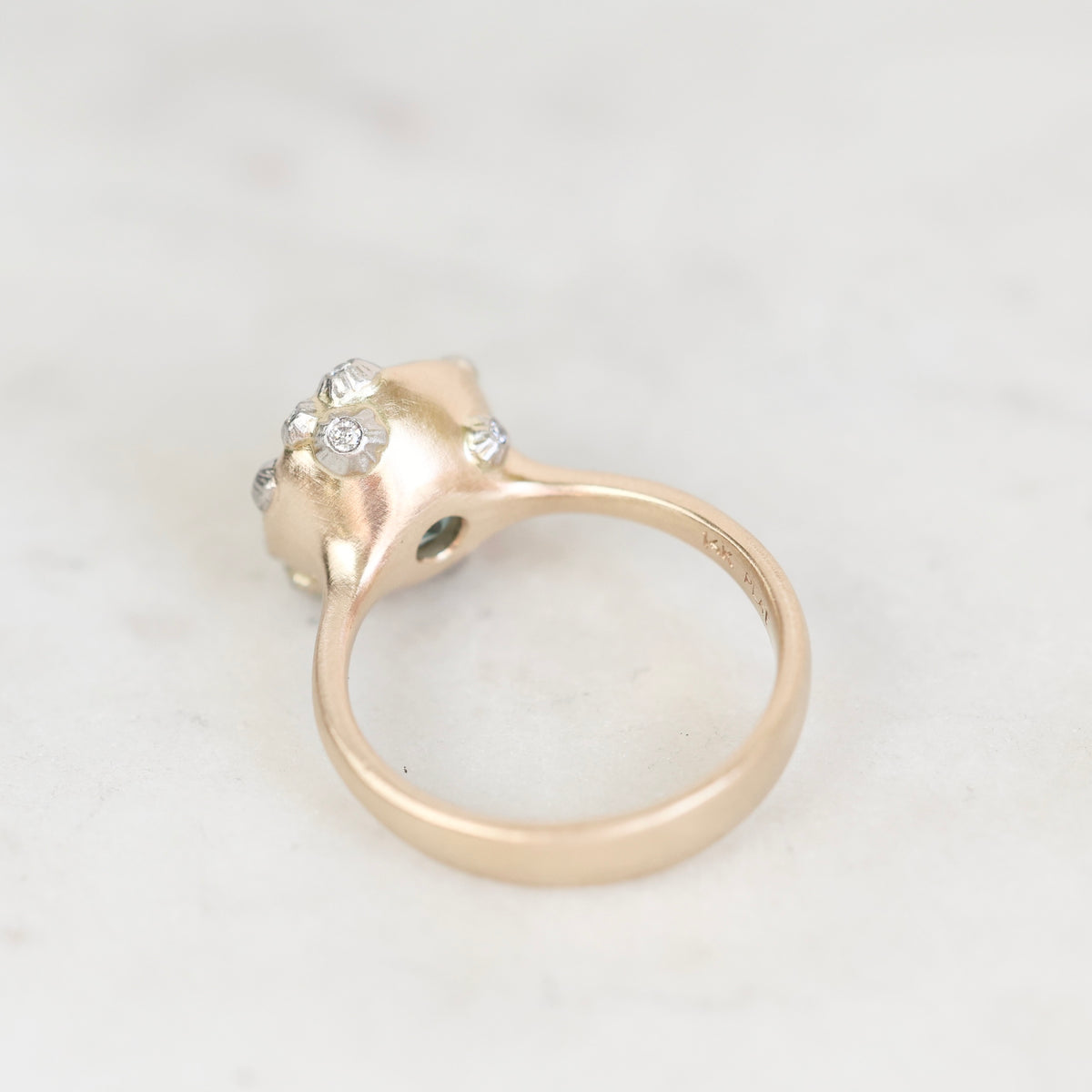 A close-up shot of a vintage yellow gold ring with a cluster of small clear gemstones on the top, set against a light marble background.