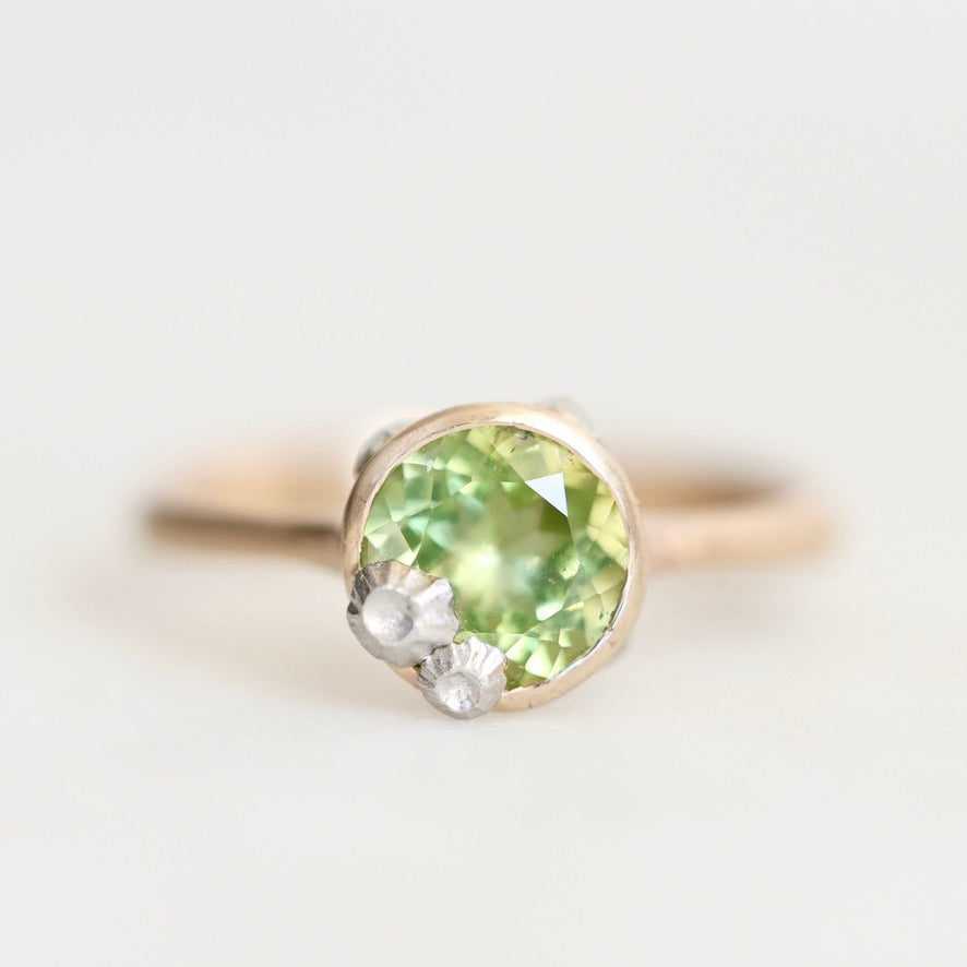 A close-up shot of a ring featuring a round-cut light green gemstone in a gold-toned setting, accented by two small white flowers on the lower right, against a soft white background.