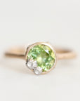 A close-up shot of a ring featuring a round-cut light green gemstone in a gold-toned setting, accented by two small white flowers on the lower right, against a soft white background.