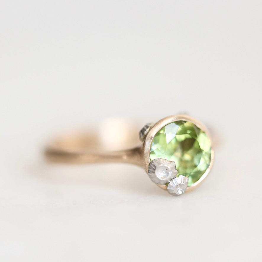 A gold ring featuring a round green peridot gemstone and two small clear accent stones, photographed on a clean white background.