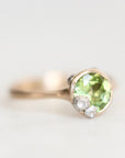 A gold ring featuring a round green peridot gemstone and two small clear accent stones, photographed on a clean white background.