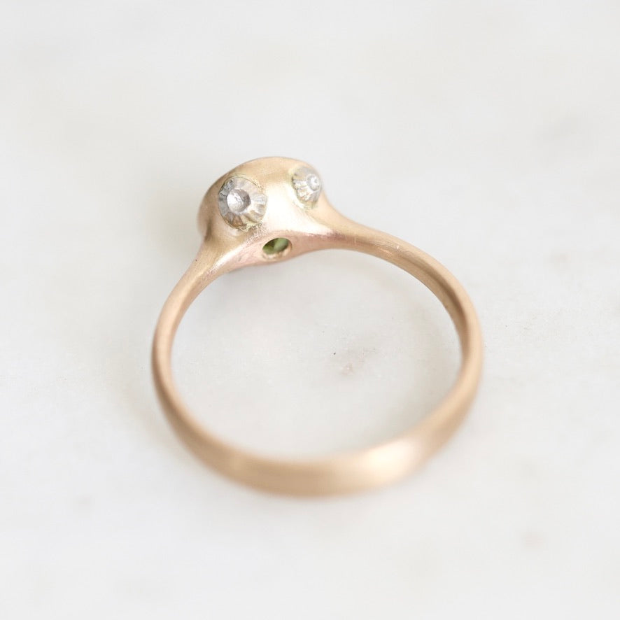 A gold ring with a unique sculptural dome top featuring two clear gemstones and a tiny green accent, photographed on a light marble surface.