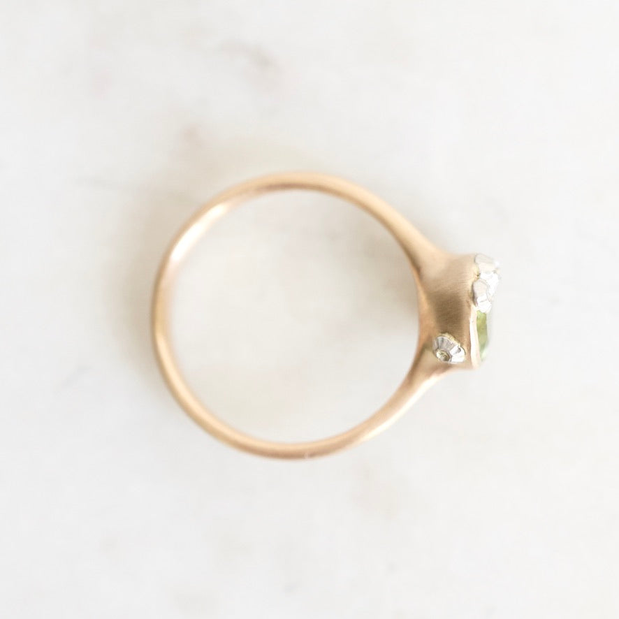 A gold ring with a small green gemstone and clear accent stones, photographed from above on a light marble surface.