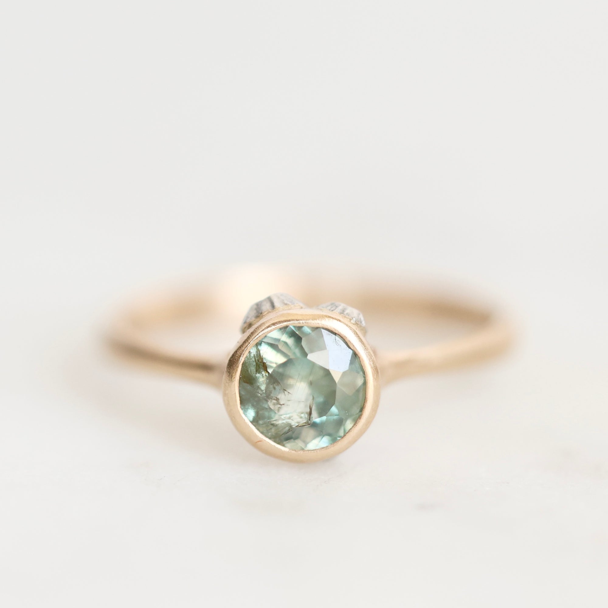 A delicate gold ring featuring a round-cut light green gemstone in a bezel setting, with small accent stones on either side of the main stone, photographed on a clean white background.