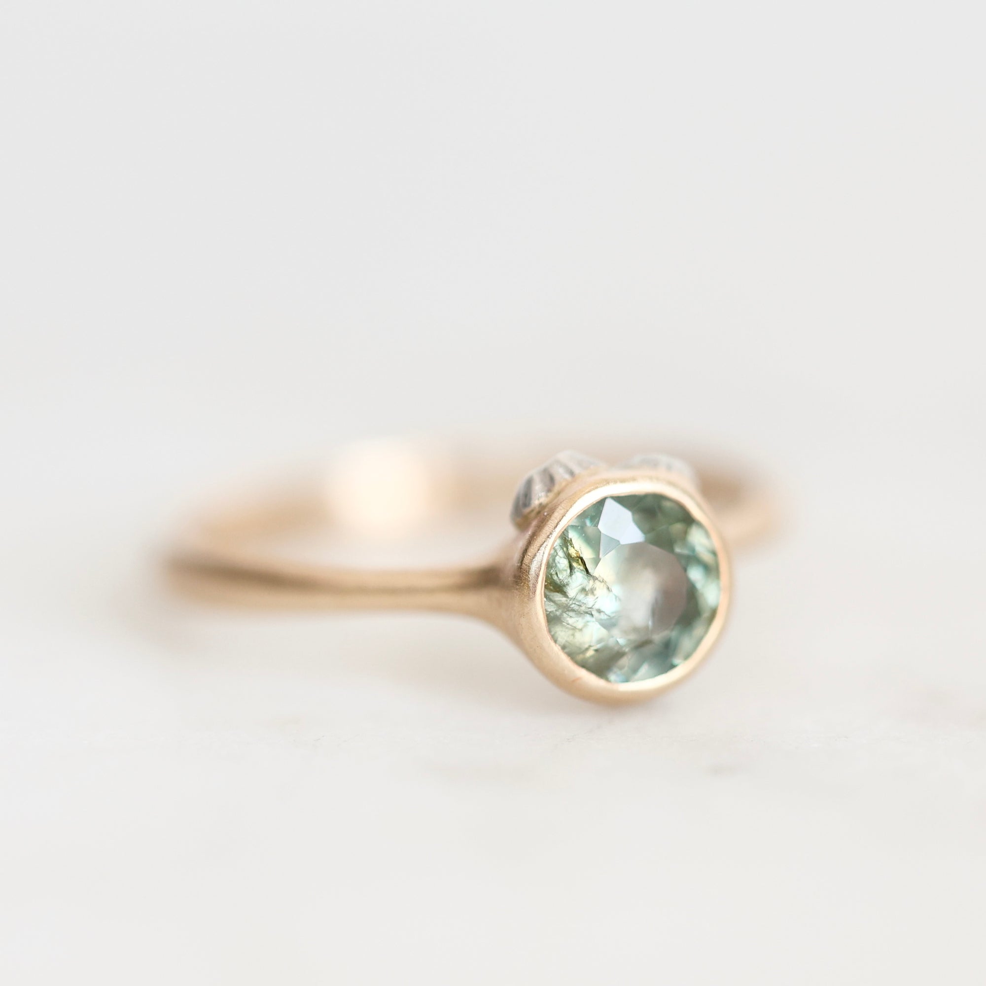 A delicate gold ring featuring a round-cut light green gemstone in a bezel setting, with a small clear accent stone on the side, photographed on a soft white background.