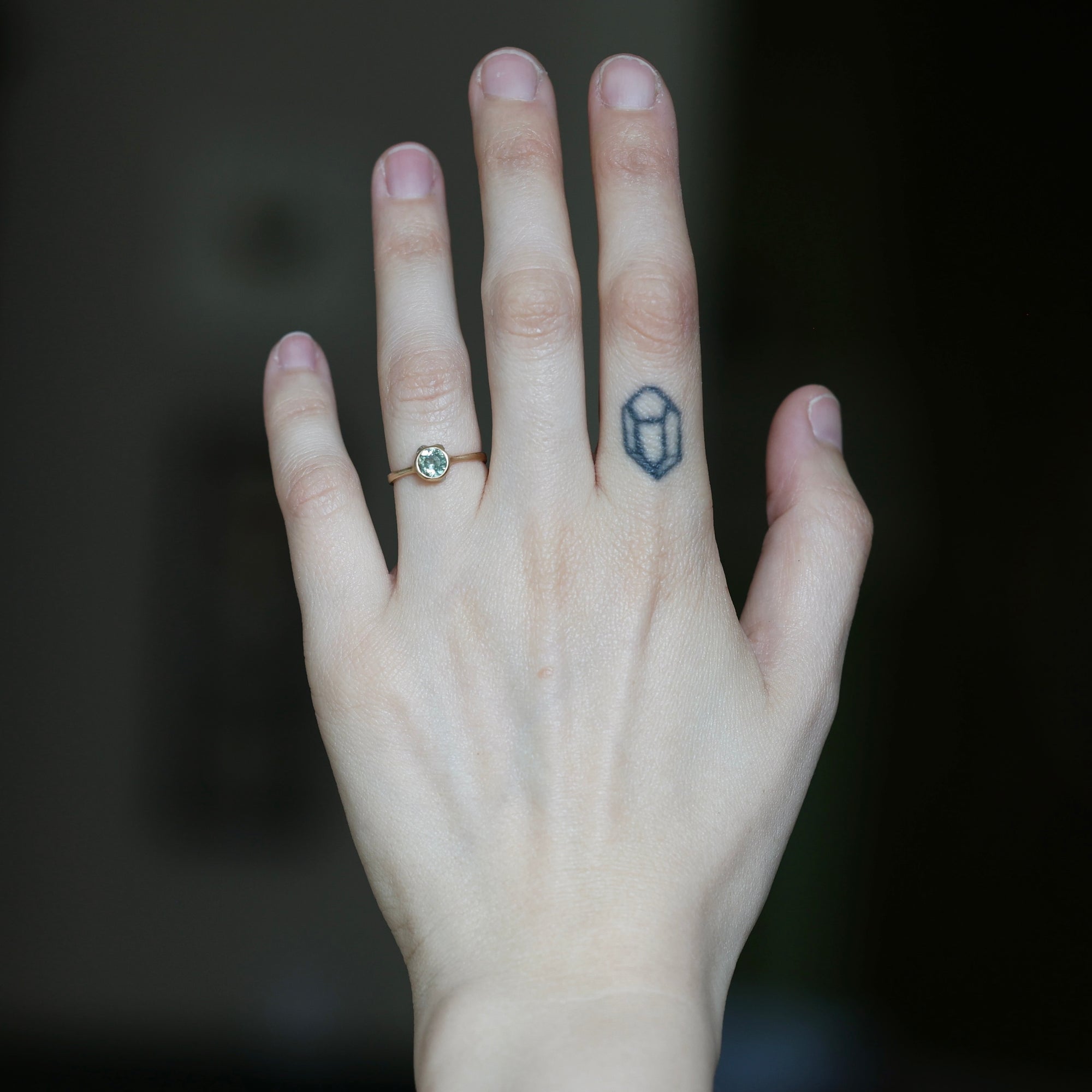 A close-up of a person's hand showing two rings: a gold band with a small round green gemstone on the ring finger, and a dark blue geometric crystal tattoo on the middle finger.