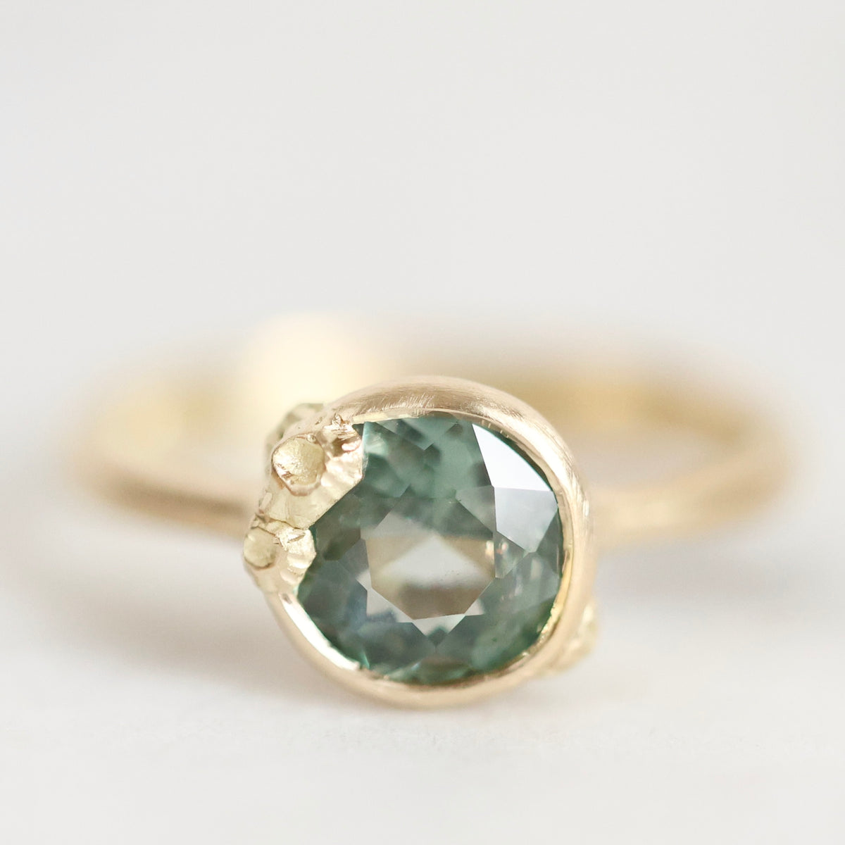 A close-up shot of a gold ring featuring a round, faceted green gemstone in a textured bezel setting, photographed against a soft, light gray background.