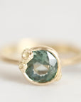 A close-up shot of a gold ring featuring a round, faceted green gemstone in a textured bezel setting, photographed against a soft, light gray background.