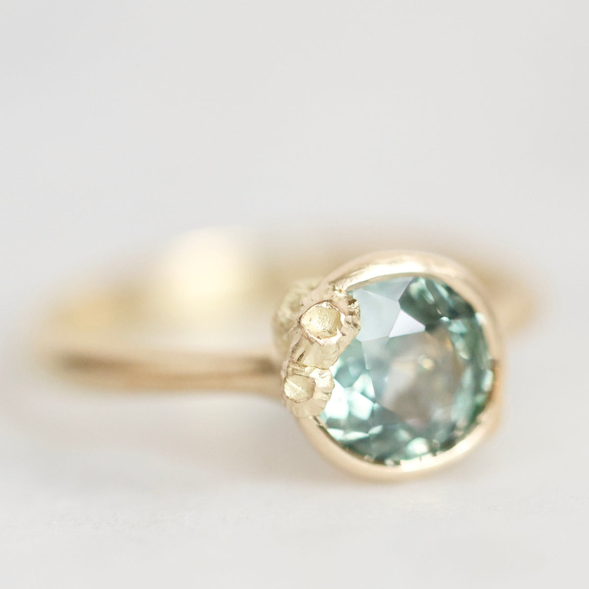 A gold ring featuring a large, round faceted light blue-green gemstone, with small clear accent stones on the band, photographed against a soft, light gray background.