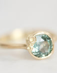 A gold ring featuring a large, round faceted light blue-green gemstone, with small clear accent stones on the band, photographed against a soft, light gray background.