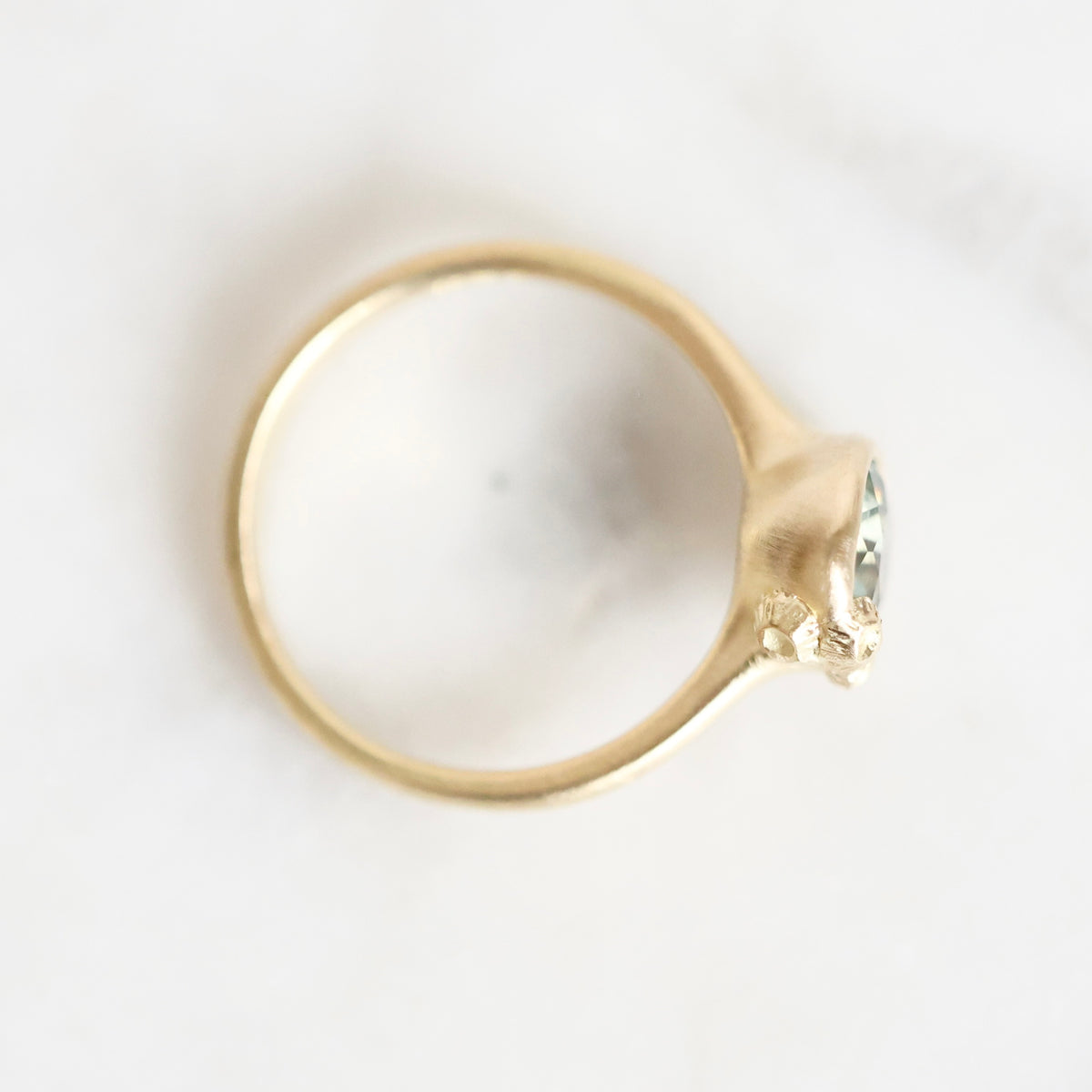 A gold ring with a small clear gemstone in a prong setting, photographed from above on a light off-white background.