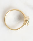 A gold ring with a small clear gemstone in a prong setting, photographed from above on a light off-white background.