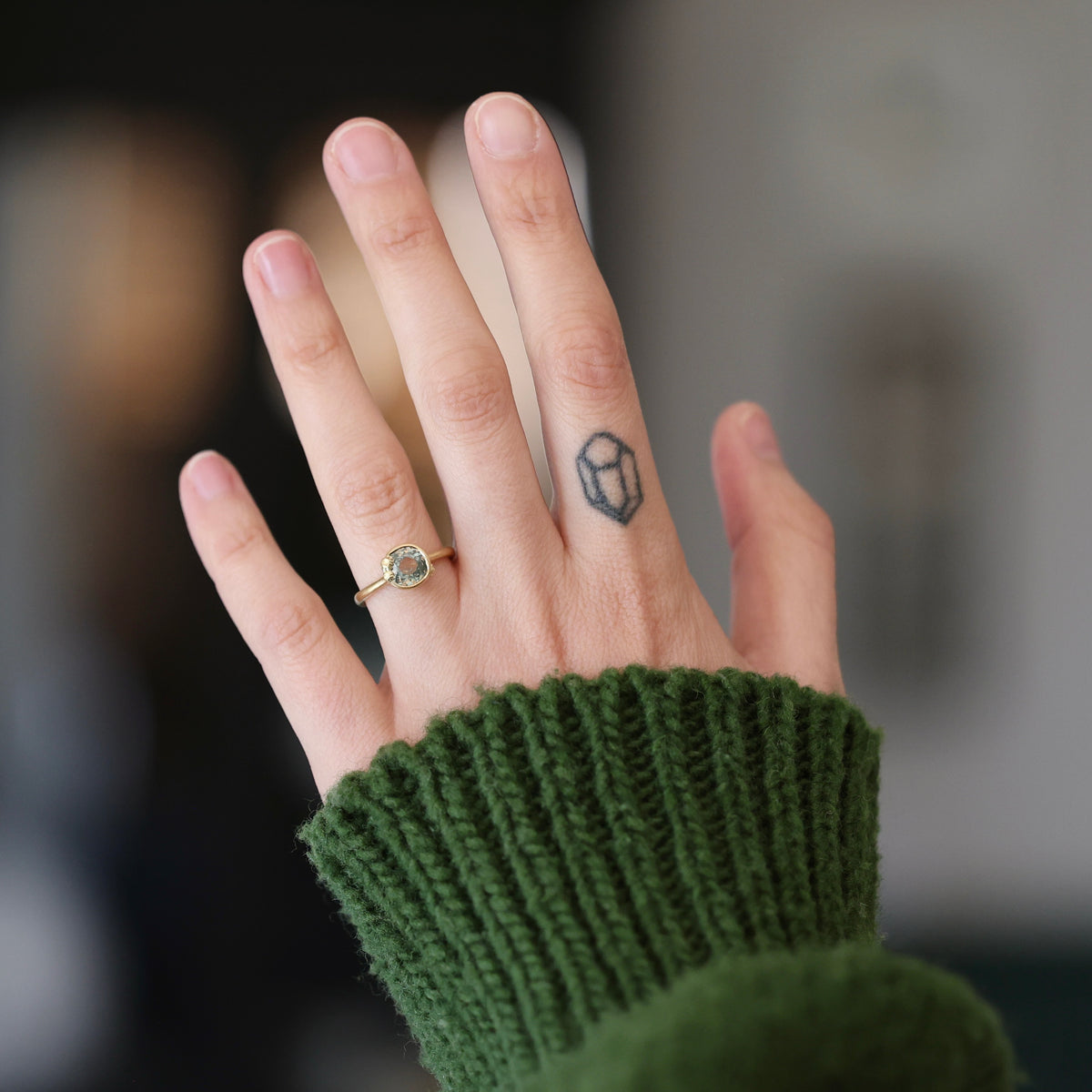 Hand with tattoo wearing gold ring with green center stone. She is wearing a green sweater against a blurred background. 