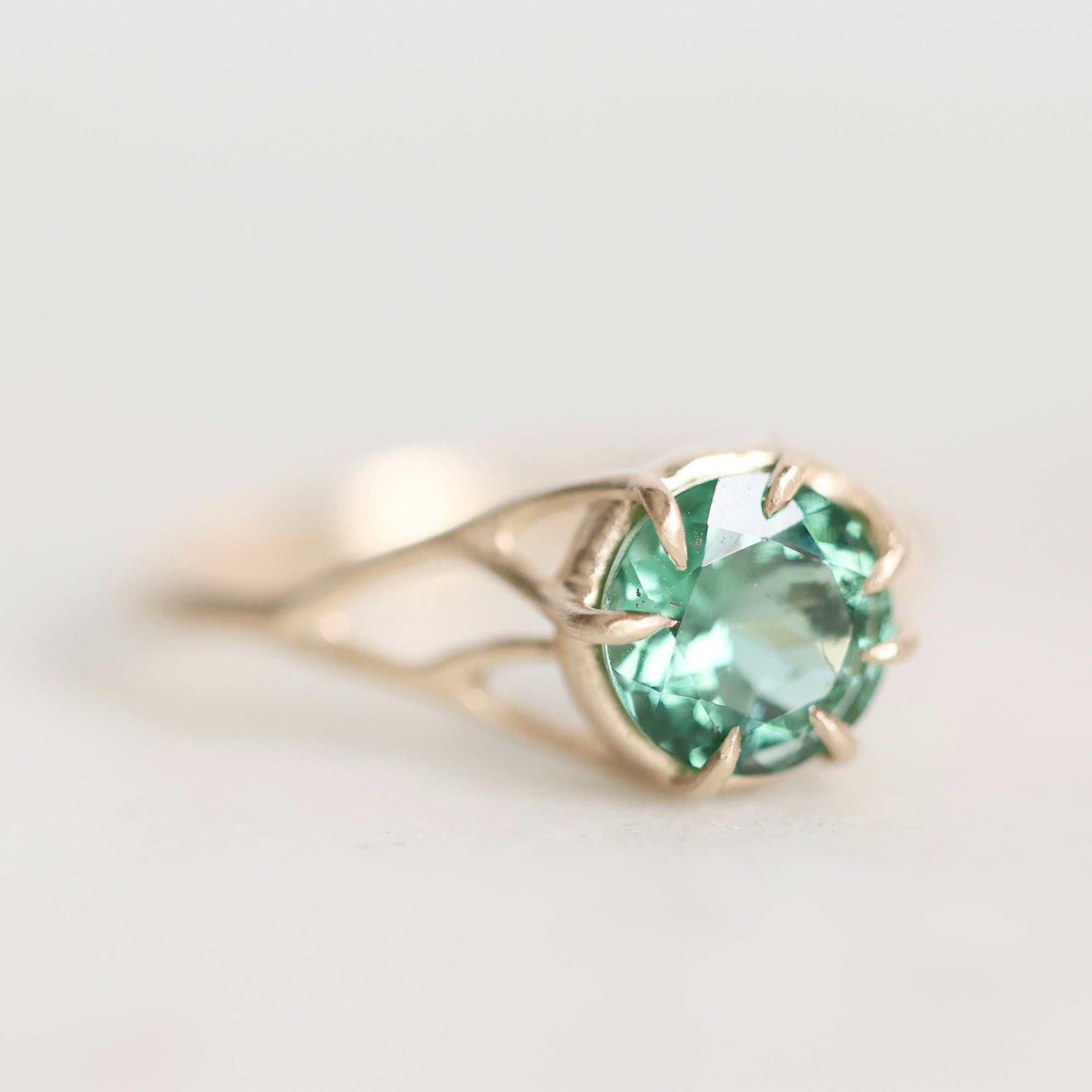 A gold ring featuring a round-cut light green gemstone in a prong setting, photographed on a clean, light gray background.