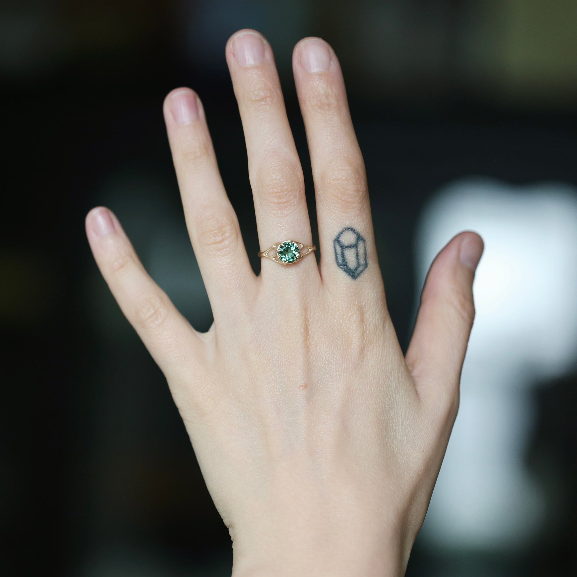 A close-up of a person's hand wearing a gold ring with a round green gemstone, set against a dark, blurred background.