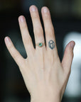 A close-up of a person's hand wearing a gold ring with a round green gemstone, set against a dark, blurred background.